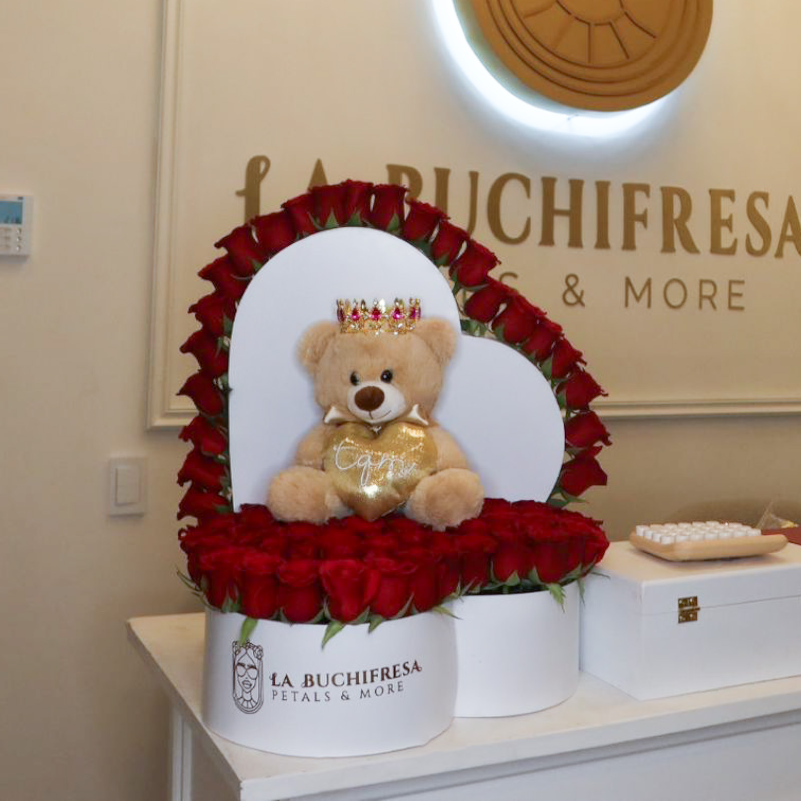 Arreglo de 100 rosas rojas premium en forma de corazon con rosas alrededor de la tapa y oso teddy de peluche con corona metalica mediana sobre caja de corazon blanca